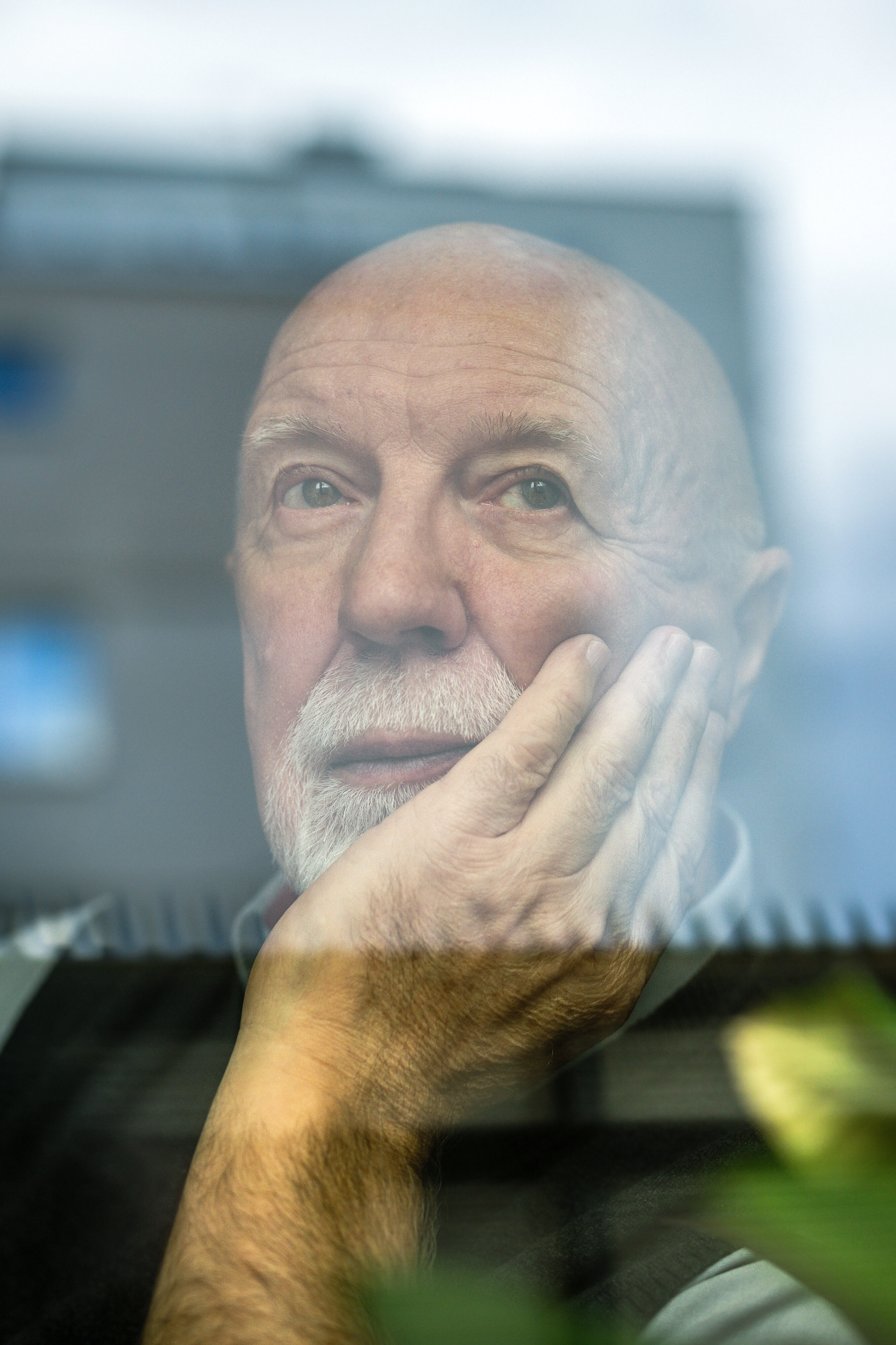 Älterer Mann sitzt nachdenklich am Fenster, stützt den Kopf mit der Hand. In der Scheibe spiegelt sich ein Hochhaus.